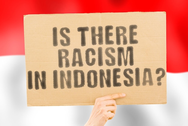 The question Is there racism in Indonesia is on a banner in men's hands with a blurred Indonesian flag in the background Interracial Issues Justice Nation Politics Peace Patriotism