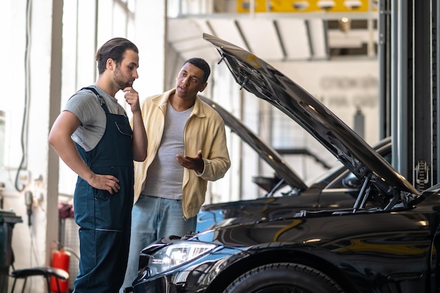 Question. Thoughtful caucasian mechanic touching chin looking into hood and dark-skinned customer asking standing near car in service