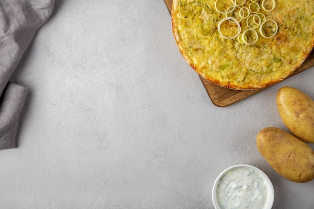 Quiche pie with leek, potatoes and cheese flat lay copy space on gray concrete background and ingredients cheese, sour cream, onion rings on round wooden cutting board.