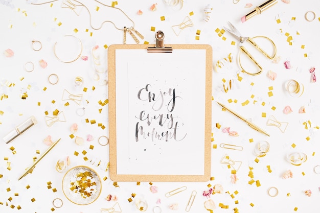 Quote Enjoy Every Moment clipboard and gold style feminine accessories pattern Golden tinsel scissors pen rings necklace bracelet on white background Flat lay top view