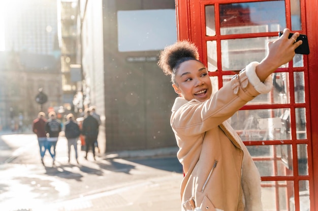 r portrait of a mixed ethnicity woman using a mobile phone and taking selfie against a red phonebox