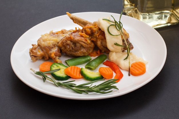 Rabbit legs baked in white wine with bechamel sauce on a ceramic plate with vegetables and rosemary. Dietary rabbit meat cooked in oven.