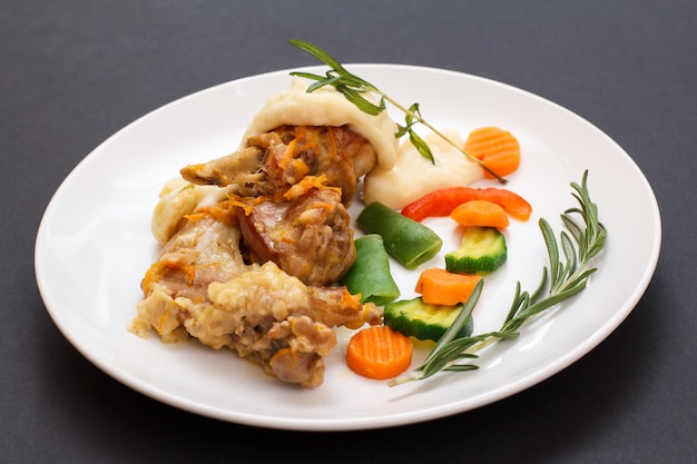 Rabbit legs baked in white wine with bechamel sauce on a white ceramic plate with vegetables and rosemary on black background. Dietary rabbit meat cooked in oven.