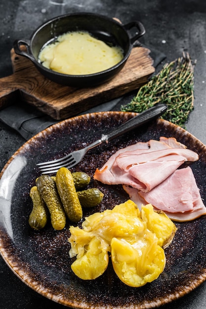 Raclette Melted cheese with boiled potato and ham on rustic plate, swiss meal. Black background. Top view.