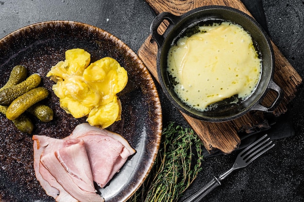 Raclette Melted cheese with boiled potato and ham on rustic plate swiss meal Black background Top view