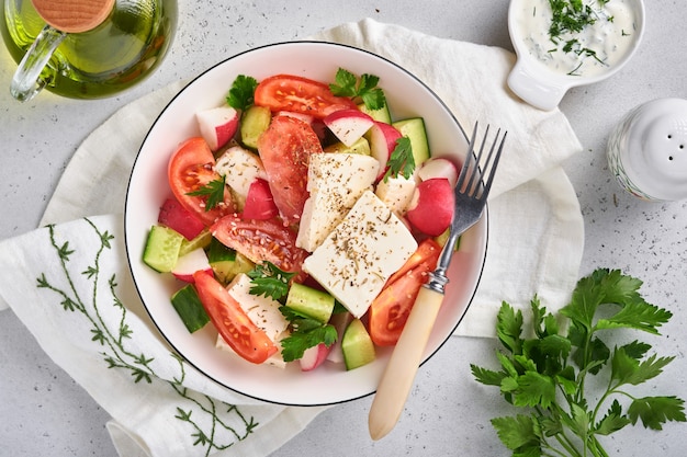 Radish, cucumber, tomato, pepper and feta cheese with spices pepper and olive oil in white bowl on grey slate, stone or concrete background. Healthy food concept. Top view.