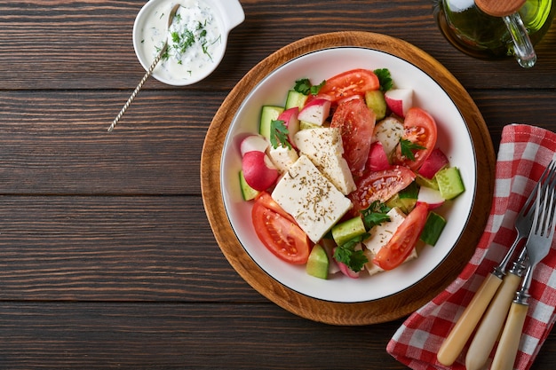 Radish, cucumber, tomato, pepper and feta cheese with spices pepper and olive oil in white bowl on old wooden dark table background. Healthy food concept. Top view.