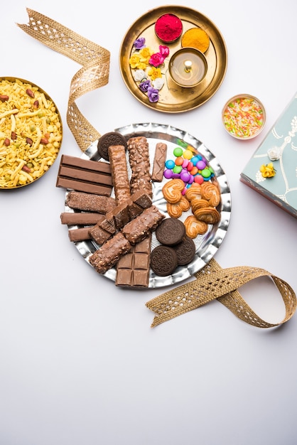 Photo raksha bandhan festival greetings: conceptual rakhi made using a plate full chocolates and biscuits with fancy band and pooja thali. selective focus