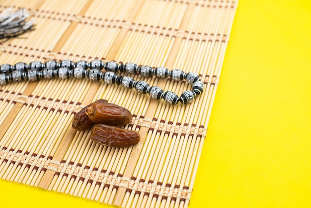 Ramadan Kareem Festival. Close up of dried dates fruit in bowl with rosary on yellow background. Copy space.