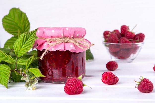 Raspberry jam in glass jar, fresh  ripe raspberry and green leaves on white wooden table