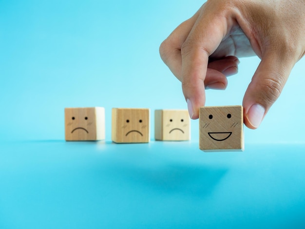 Rating customer service evaluation feedback and satisfaction survey concepts Hand chosen one from others choice holding happy smile emoticon face on the wooden cube block on blue background