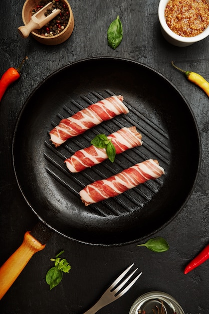 Raw bacon rolls in a grill frying pan with herbs, fork and mustard. Top view closeup.