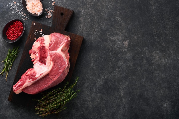 Raw beef meat Rough piece of meat on bone for roast or soup with salt pepper thyme and rosemary on a black concrete background Entrecote Raw cowboy steak Top view