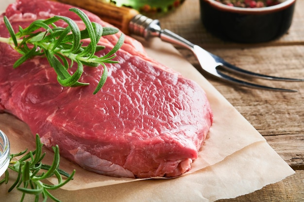 Raw beef steak Fresh beef rib eye steak with fork rosemary salt and pepper on piece of parchment paper on old wooden background Top view Mockup for design idea