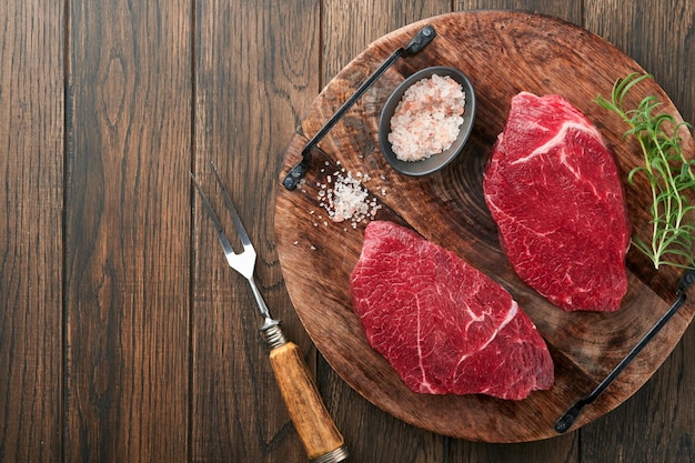 Raw beef steak Marbled raw fresh Ribeye steak with rosemary salt and pepper on cutting board on dark concrete background Raw beef steak and spices for cooking