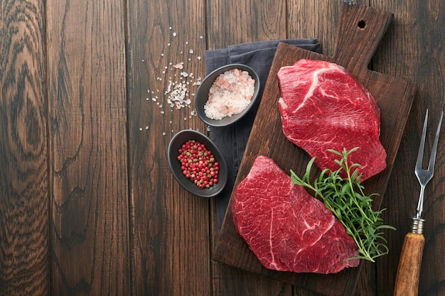 Raw beef steak Marbled raw fresh Ribeye steak with rosemary salt and pepper on cutting board on dark concrete background Raw beef steak and spices for cooking