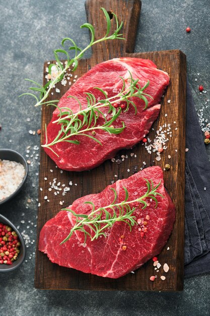 Raw beef steak Marbled raw fresh Ribeye steak with rosemary salt and pepper on cutting board on dark concrete background Raw beef steak and spices for cooking