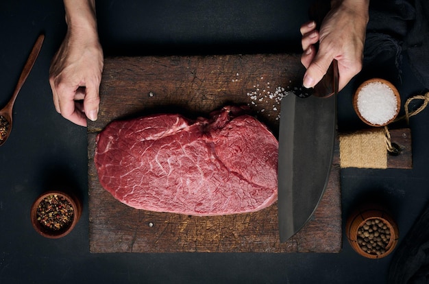 Raw beef tenderloin lies on a wooden cutting board and spices for cooking on a black table top view A woman's hand holds a large kitchen knife in her hand