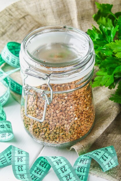 Raw buckwheat in a glass jar with a centimeter on sacking on a wooden table background of parsley.