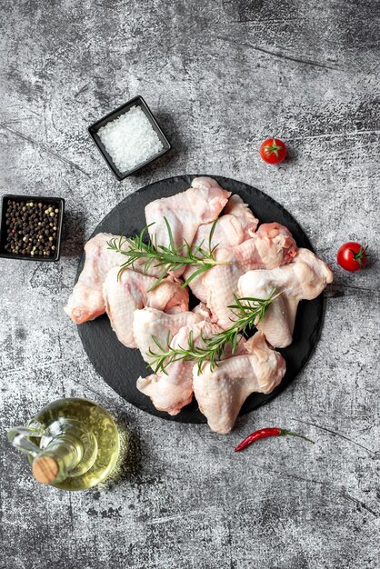 Raw chicken wings on a black plate with spices and herbs on a gray background.