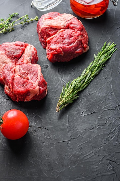 Raw chuck eye roll steak cuts with rosemary spicy chili oil and peppercorns on black textured background side view with space for text