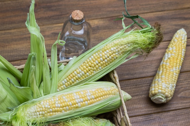 Raw corn cobs with corn silk and leaves in wicker basket. Water bottle. Wooden background. Flat lay