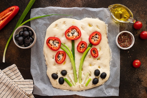 Raw focaccia decorated with vegetables on dark table with ingredients Garden focaccia