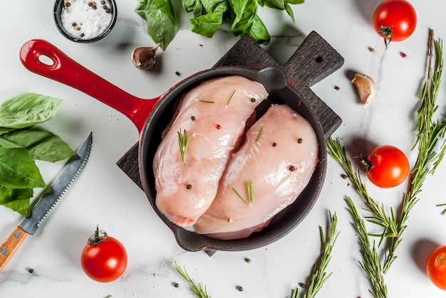 Raw fresh chicken breast fillet with herbs and spices for cooking, in iron cast skillet, white marble table, copy space top view