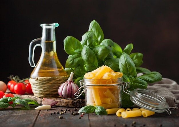 Raw fresh penne pasta in glass jar with oil and garlic basil plant on wooden table background Macro