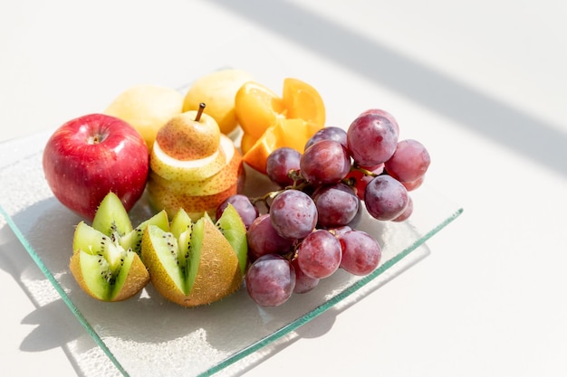 Raw fruits berries platter oranges kiwi grapefruit grapes pear apples on the plate on the white tabl