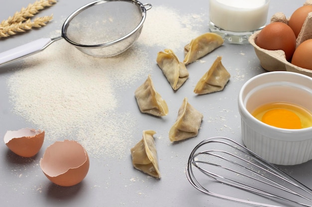 Raw gyoza flour and sieve on table Broken egg in bowl whisk and eggshell Top view Gray background