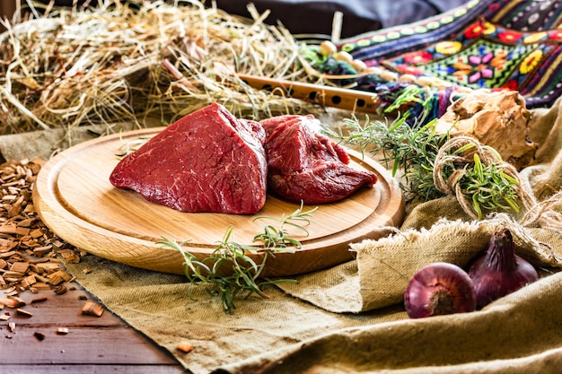 Raw juicy meat on a round wood board. Near rural flute, hay and shepherd's bag