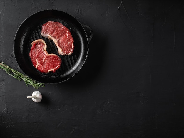 Raw juicy steaks in an iron grilling pan black background Fresh meat garlic green twig Cooking restaurant concept Closeup shot Top view