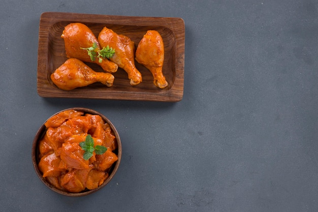 Raw marinated chicken cubes and legs arranged in a wooden tableware garnished with fresh coriander and mint leaf which is placed on grey textured background.