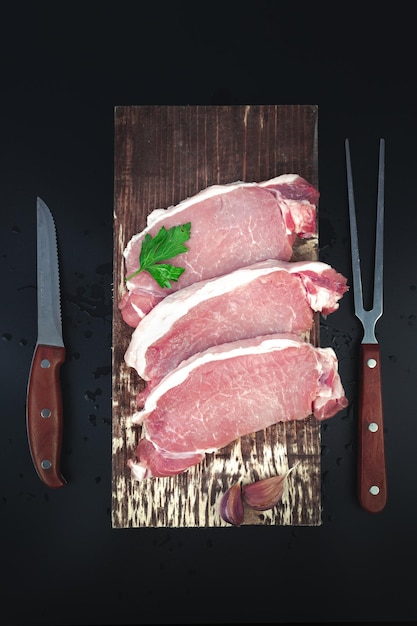 Raw meat steaks with ingredients for cooking knife and meat fork on dark background