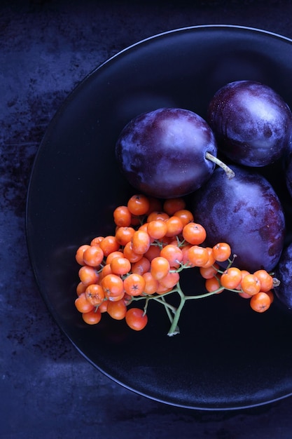 Raw and organic fruits harvest from garden closeup Ripe juicy homemade plums and rowanberries on a black plate on a dark background