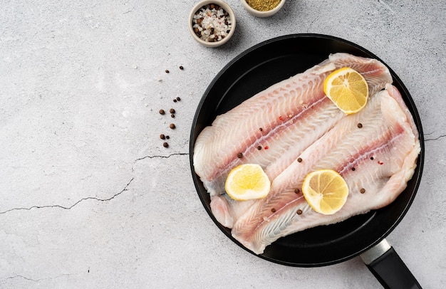 Raw pangasius fish fillet with lemon and spice in frying pan on concrete background