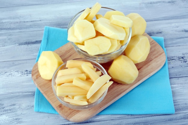 Raw peeled and sliced potatoes in glass bowls on cutting board on color wooden background