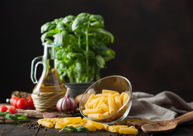 Raw penne pasta in glass bowl with oil and garlic basil plant and tomatoes with pepper and linen towel on wooden table Space for text