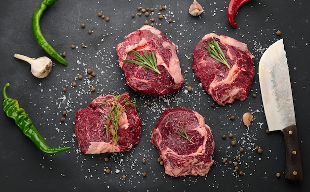 Raw piece of beef ribeye with rosemary, thyme on a black table, top view