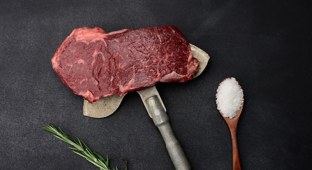 Raw piece of beef steak lie on an iron knife, black surface. Classic steak, top view