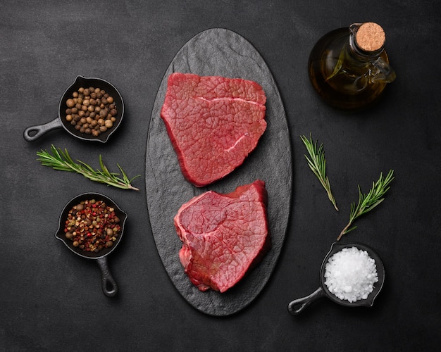 Raw piece of beef with spices pepper rosemary sprig salt and olive oil on a wooden board black background