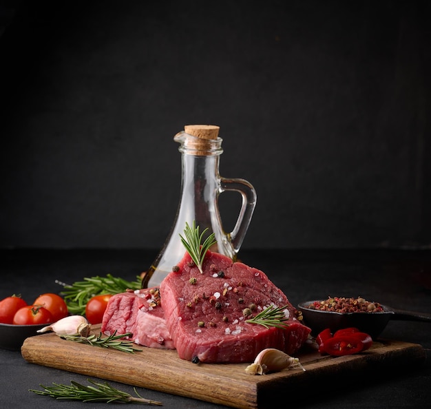 Raw piece of beef with spices pepper rosemary sprig salt and olive oil on a wooden board black background