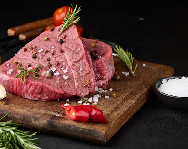 Raw piece of beef with spices pepper rosemary sprig salt and olive oil on a wooden board black background