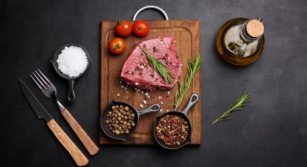 Raw piece of beef with spices pepper rosemary sprig salt and olive oil on a wooden board black background