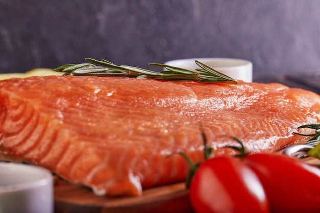 Raw salmon fillet pepper salt dill lemon rosemary and cherry tomatoes on a dark wooden table.