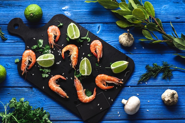 Raw shrimp on a blue background lie on a black cutting board next to lime greens , mushrooms