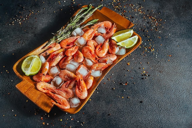 Photo raw shrimp with ice, lime and rosemary sprigs lie on a wooden tray with seasonings, dark background