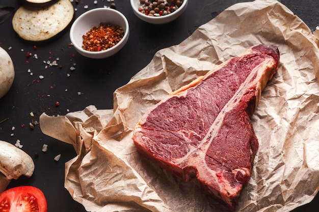 Raw t-bone steak on craft paper on dark background. Prime meat on black table with spices, copy space