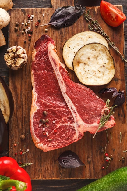 Raw t-bone steak. Fresh meat, rosemary, vegetables on wooden board at black background. Organic ingredients for restaurant meals, copy space, top view